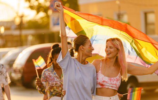 Young lesbian couple smiling and walking with rainbow flag during pride parade at city street